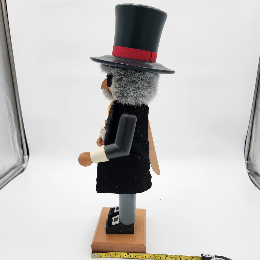 A Christian Ulbricht nutcracker decorative figure shown from the side on a white studio background, wearing a black top hat with a red band, black coat, gray wings, and standing on a wooden base. A measuring tape is visible along the bottom edge.