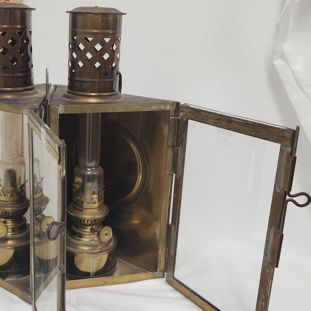 A vintage brass oil lantern with glass doors swung open, showing an antique brass oil lamp inside. The lantern features a cutout metal top and a dark metal frame, photographed against a white studio background.