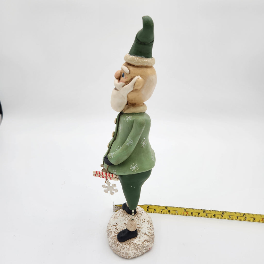 A small Janell Berryman Pumpkinseeds "Sage and Peppermint Santagurine shown in profile on a white studio background, with a yellow tape measure placed along the base to indicate size. The figurine wears a green hat and green jacket with white snowflake details, has a beige face and beard, and stands on a textured beige base.