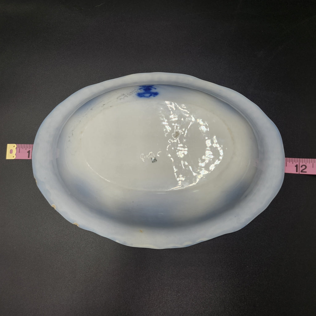 An oval white ceramic plate with a blue painted design near the rim, shown on a dark background with pink measuring tapes on both sides to indicate size. 