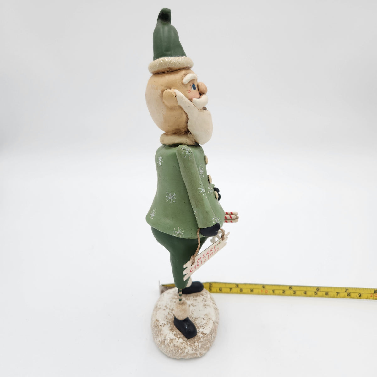 A handcrafted   Janell Berryman Pumpkinseeds "Sage and Peppermint Santa from the side on a white studio background, with a yellow tape measure placed along its base to indicate scale.