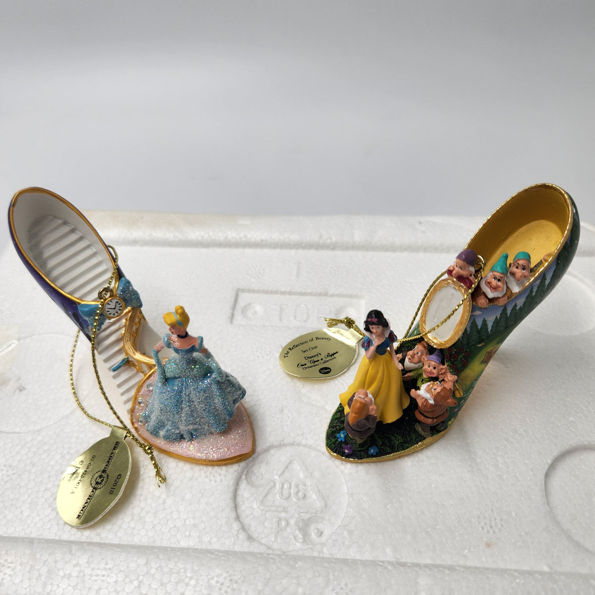 A pair of Disney-themed shoe-shaped ornaments displayed on white foam packaging. One ornament features Cinderella in a blue ball gown on a pink base, and the other features Snow White in a yellow dress with the Seven Dwarfs on a green base.