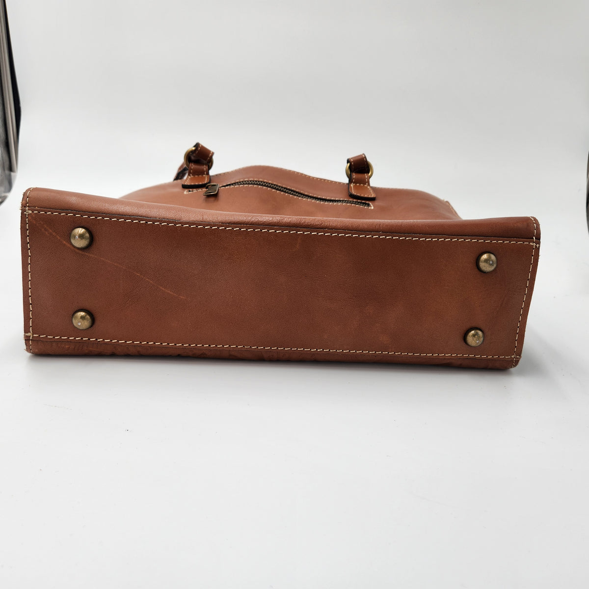 A brown leather handbag shown from the bottom front angle, featuring brass-tone rivets on the bottom corners, white contrast stitching, and a zipper closure at the top, photographed on a white studio background.