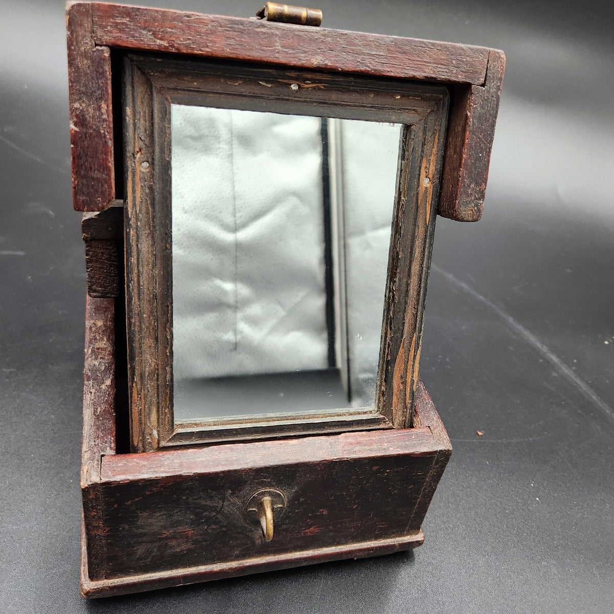 An antique wooden wall-mounted mirror with a dark brown wood frame and brass hardware, shown on a dark gray surface.