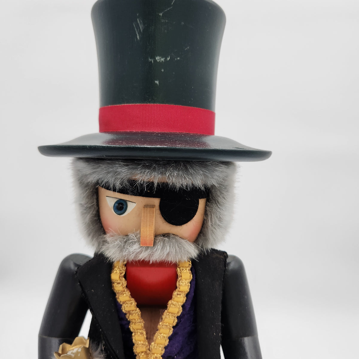 A close-up of a wooden pirate nutcracker wearing a tall black top hat with a red band, a black eyepatch, a gray faux-fur hat and beard, a black jacket, and a gold chain necklace.