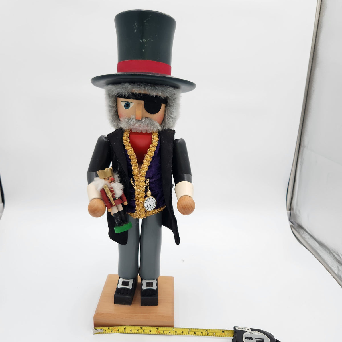 A decorative wooden nutcracker figurine standing on a small wooden base against a white studio background. The figure wears a tall black top hat with a red band, a black coat over a red shirt, a dark purple vest with a large gold chain necklace, gray pants, and black shoes.Christian Ulbrickt