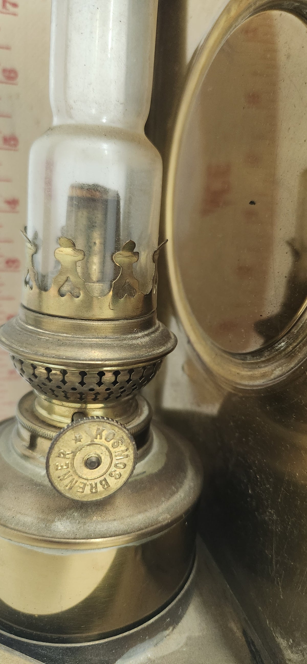A vintage brass oil lamp with a clear glass chimney and a brass keyhole-shaped adjustment knob, shown next to a round brass object on a tabletop.