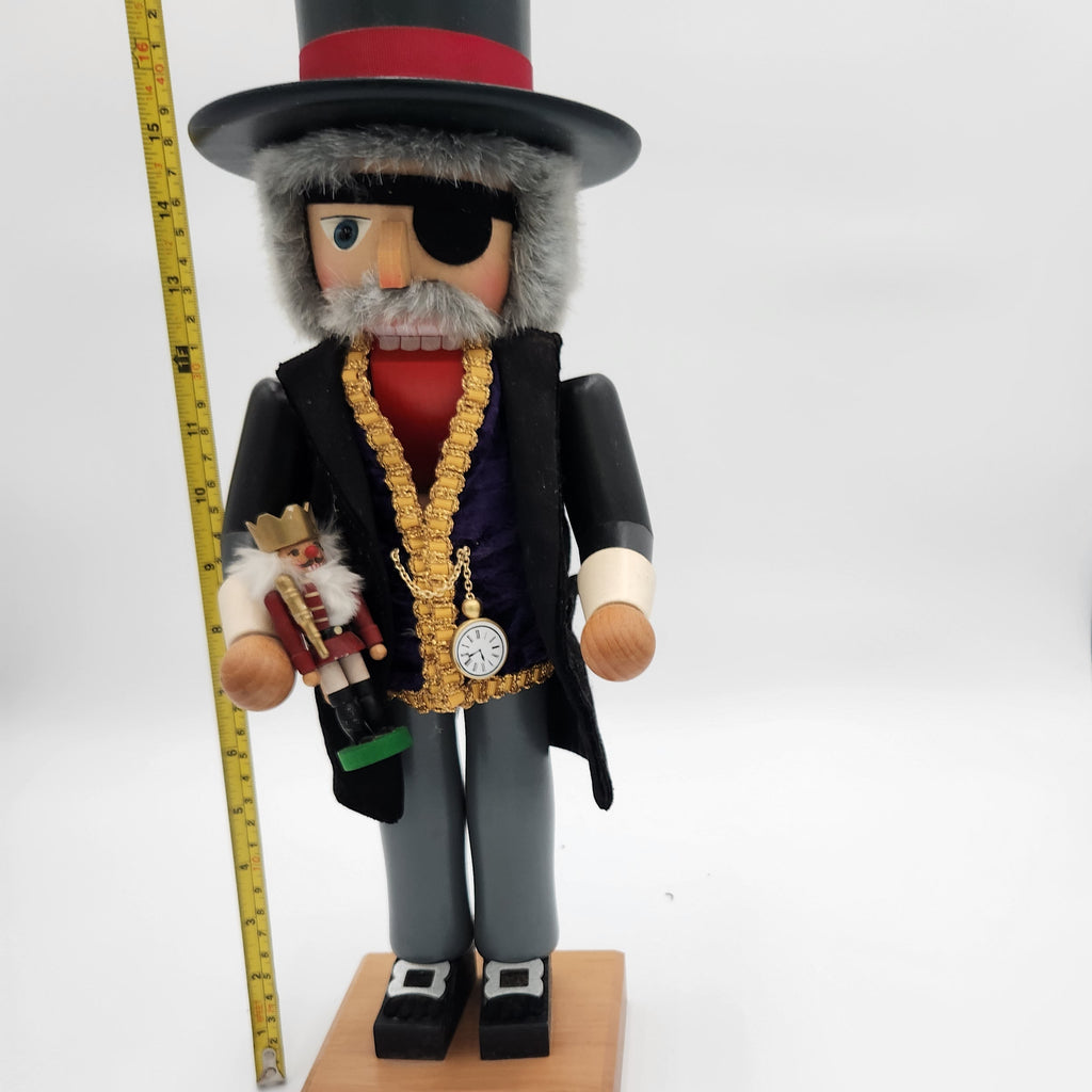 A decorative wooden nutcracker figurine wearing a black top hat with a red band, black coat, gray pants, and a gold chain necklace with a pocket watch pendant. It stands on a small wooden base next to a yellow measuring tape against a white studio background.Christian Ulbrickt