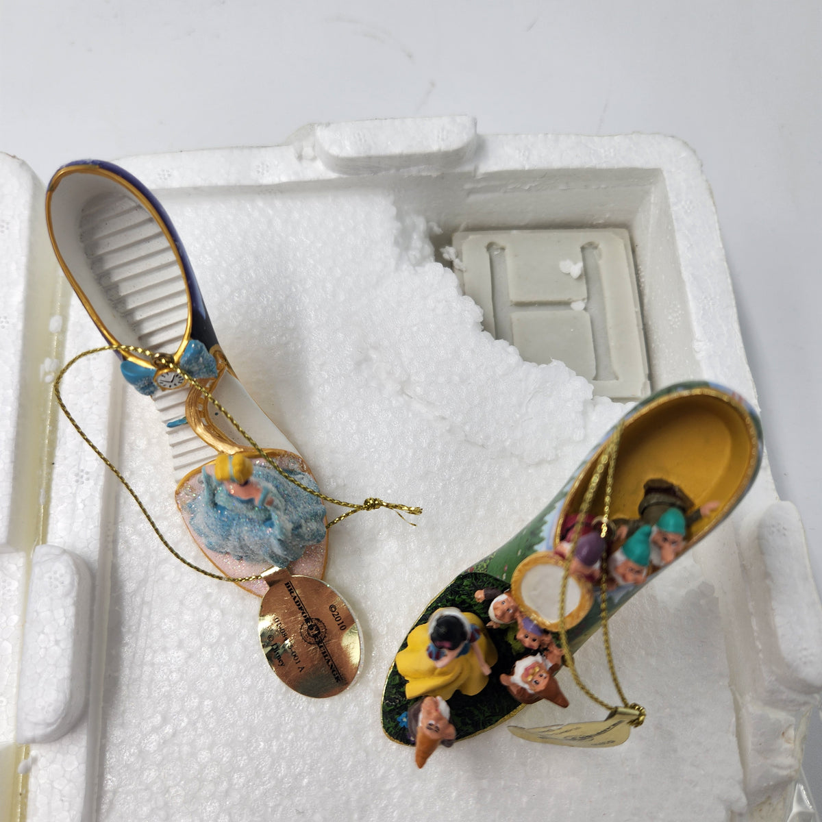 A pair of decorative Cinderella-themed glass slipper ornaments displayed inside their white foam packaging. One slipper features a blue and gold exterior with a hanging ornament of Cinderella in a blue gown, and the other features a green and gold exterior with a hanging ornament of Cinderella in a yellow gown. Cinderella Snow White