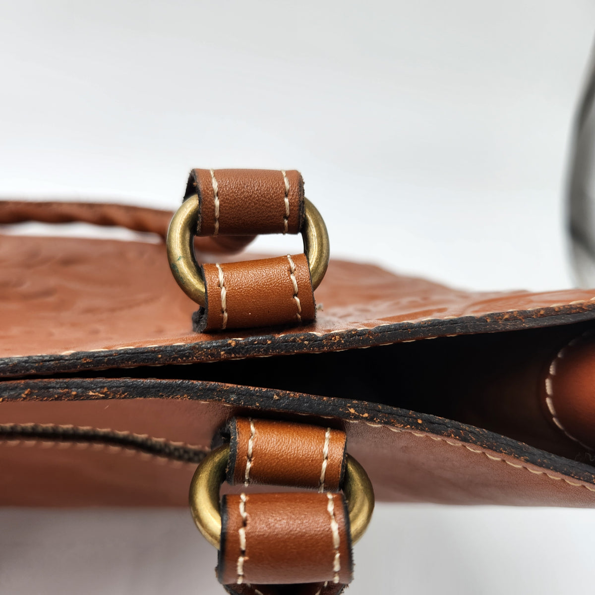 Close-up of a brown leather bag showing a strap with white stitching, brass-toned metal rings, and a textured leather surface.