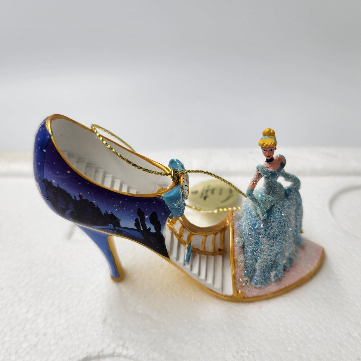 A decorative miniature high-heeled shoe ornament styled like Cinderella's glass slipper, with a blue and gold exterior, a small figure of Cinderella in a glittery blue gown on the heel, and a staircase design on the side, photographed on white foam packaging.
