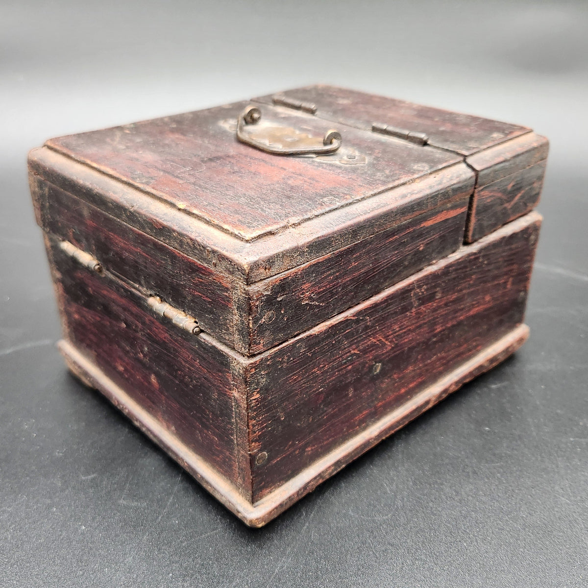 A small vintage wooden box with a dark reddish-brown finish and brass latch, photographed on a dark gray surface.