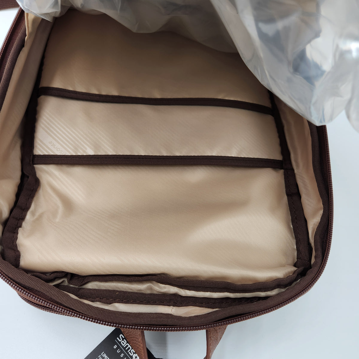 Open view of a brown and beige backpack showing the interior lining, zip compartments, and a plastic protective cover partially peeled back. A small visible hangtag shows the brand Samsonite.