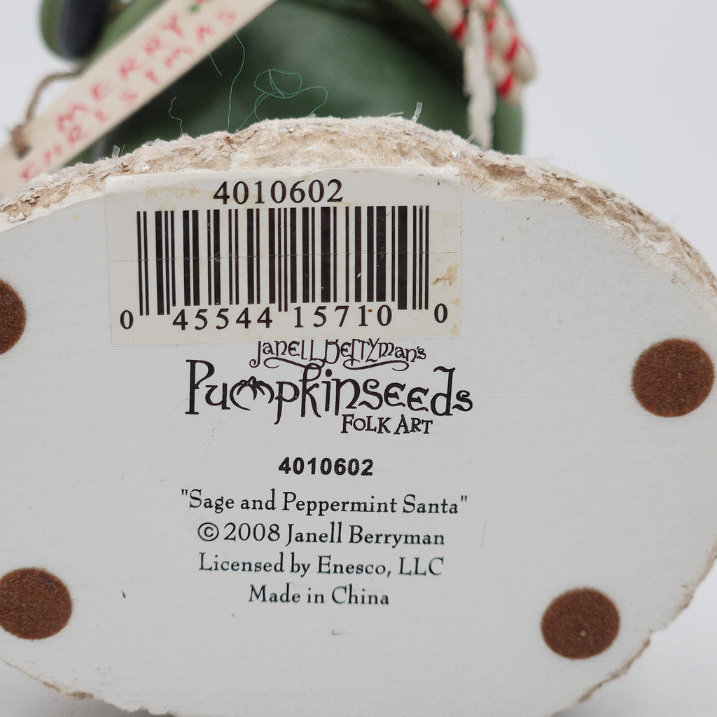 Close-up of the bottom of a decorative pumpkin seed ornament showing a barcode label, product text for 'Pumpkinseeds Folk Art' and 'Sage and Peppermint Santa', plus part of the hanging string and green top visible against a white background.