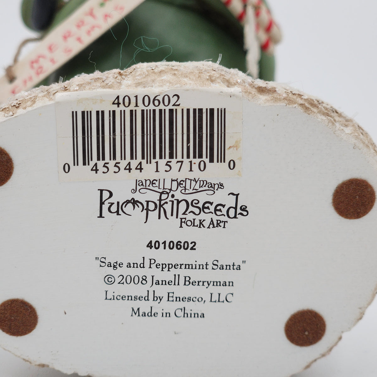 Close-up of the bottom of a decorative pumpkin seed ornament showing a barcode label, product text for 'Pumpkinseeds Folk Art' and 'Sage and Peppermint Santa', plus part of the hanging string and green top visible against a white background.