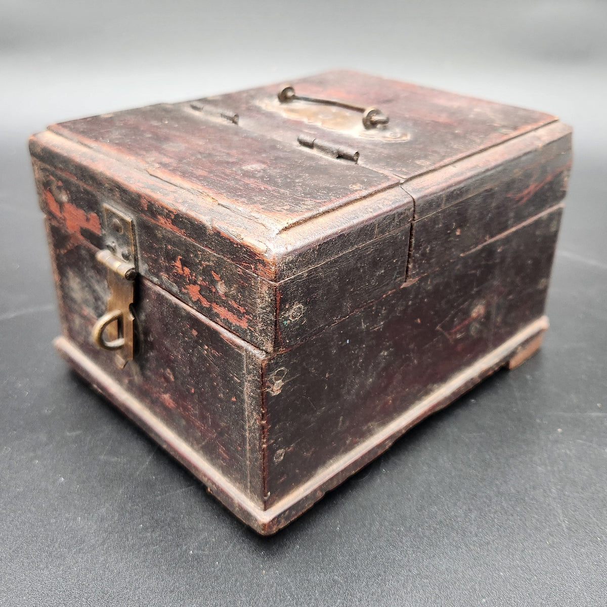 An antique dark brown wooden box with a weathered finish and brass latch, photographed on a dark gray background.