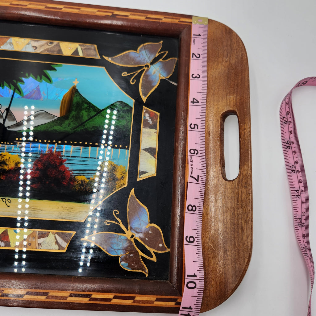 A close-up of a rectangular wooden serving tray with a colorful hand-painted landscape panel and butterfly motifs, shown with a pink measuring tape along its side on a white background. Vintage Brazilian Butterfly Wing Marquetry Tray Rio De Janeiro Inlaid Wood Label