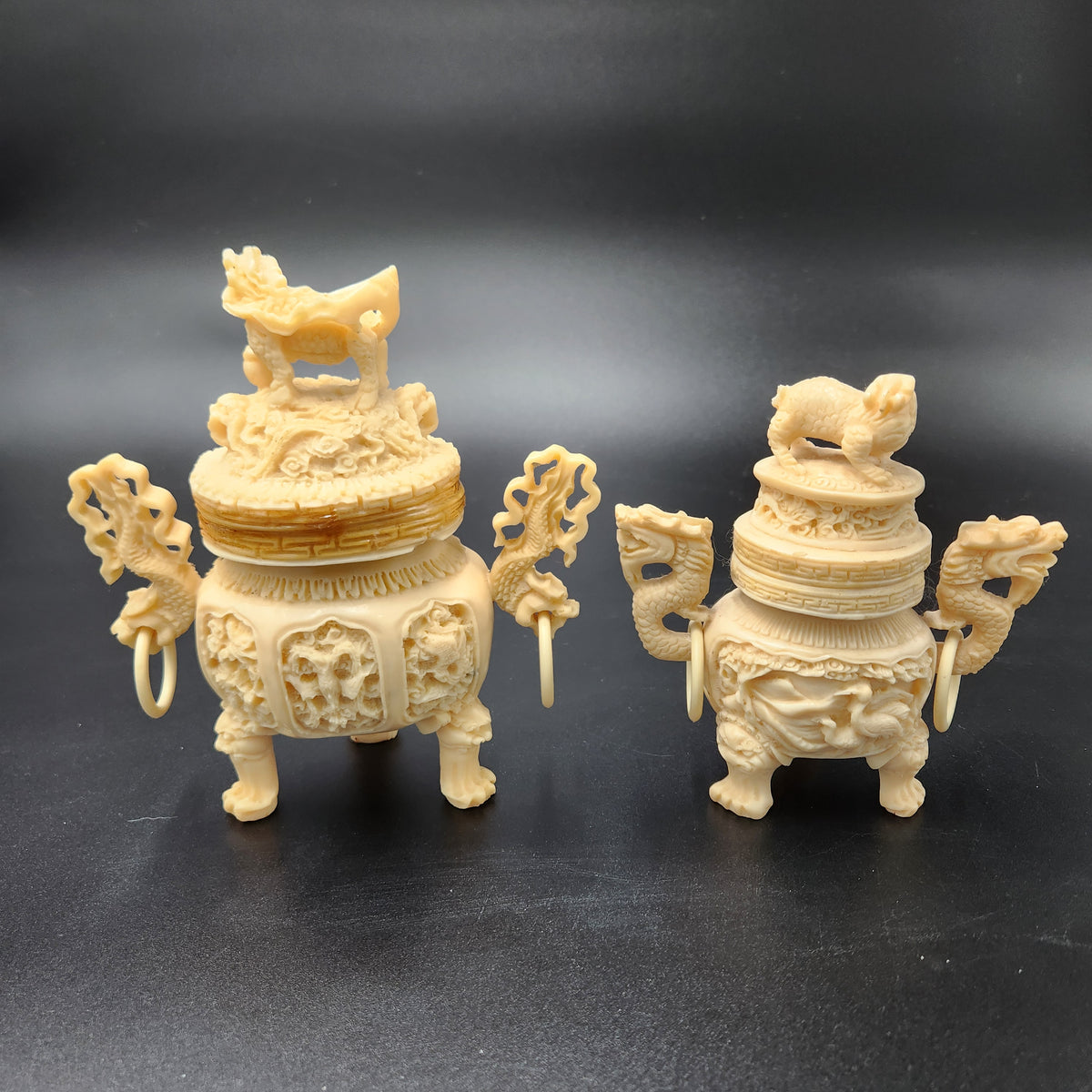 Two ornate cream-colored carved Chinese bronze-style incense burners with dragon handles and lion capitals, displayed on a dark gray background. Vintage Chinese Carved Resin Faux Ivory Incense Burners Tripod Censers 