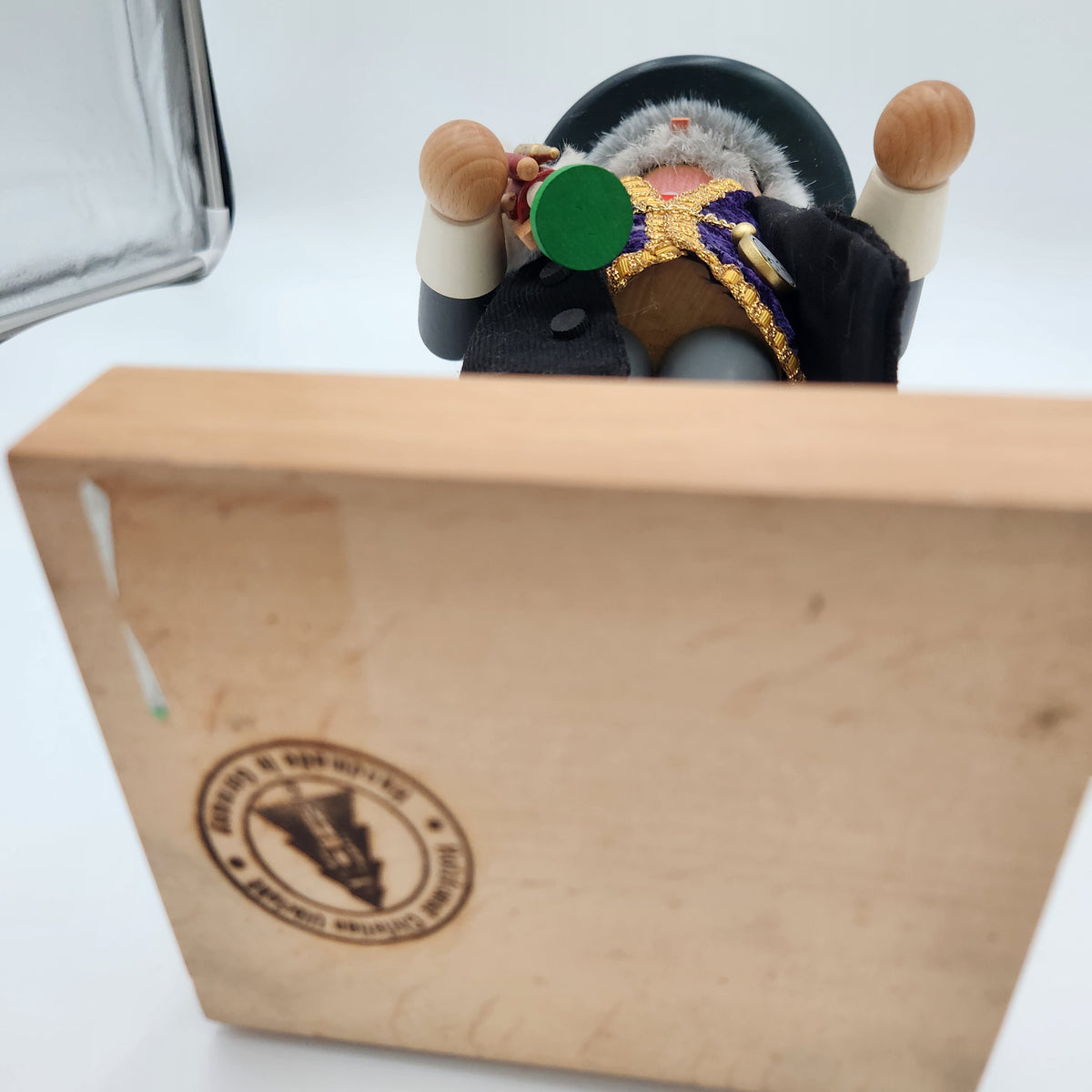 A close-up of a nutcracker figure positioned behind a large light wood box. The nutcracker has wooden arms with light brown knobs, a black coat, a purple and gold embroidered chest piece, and a gray beard.