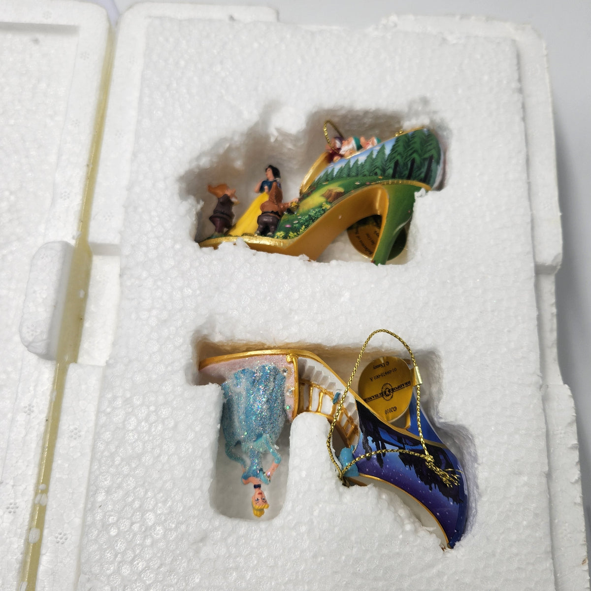 Three hand-painted glass high-heeled shoes displayed in a white foam insert box. The top shoe is green with a gold heel and a fairy illustration; the bottom-left is light blue with a gold heel and a princess illustration; the bottom-right is dark blue with a gold heel and a mermaid illustration. Cinderella Snow White