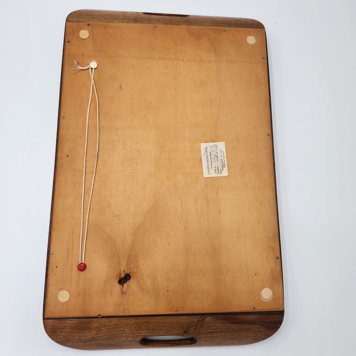 The underside of a wooden serving tray or board, showing a light natural wood grain surface with a dark brown wooden rim, corner feet with circular pads, a hanging cord with a red bead, and a small paper label. Vintage Brazilian Butterfly Wing Marquetry Tray Rio De Janeiro Inlaid Wood Label