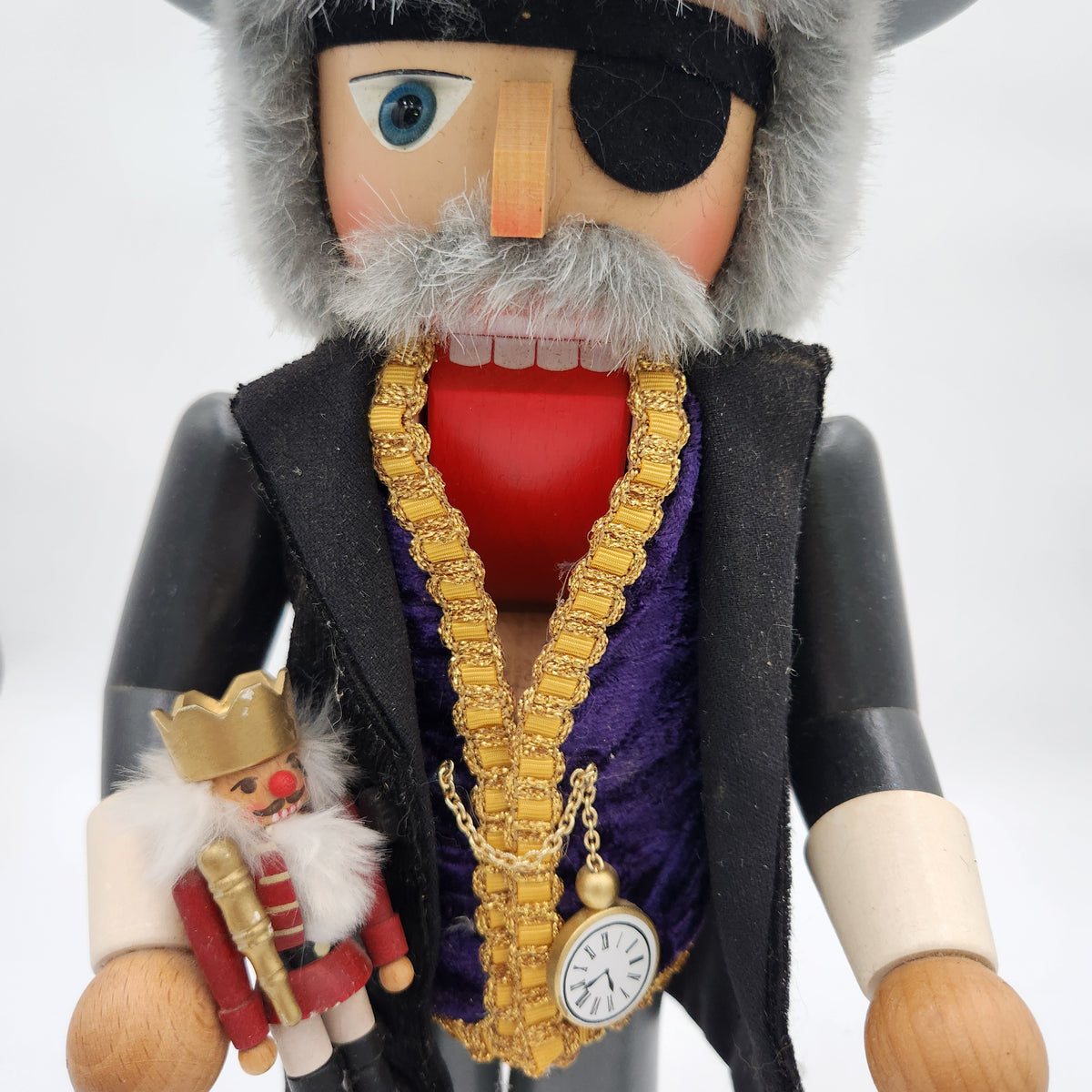 A close-up of a nutcracker figurine with a gray beard, black eyepatch, red shirt, black coat, purple vest, and gold chain accessories, holding a small miniature nutcracker.
