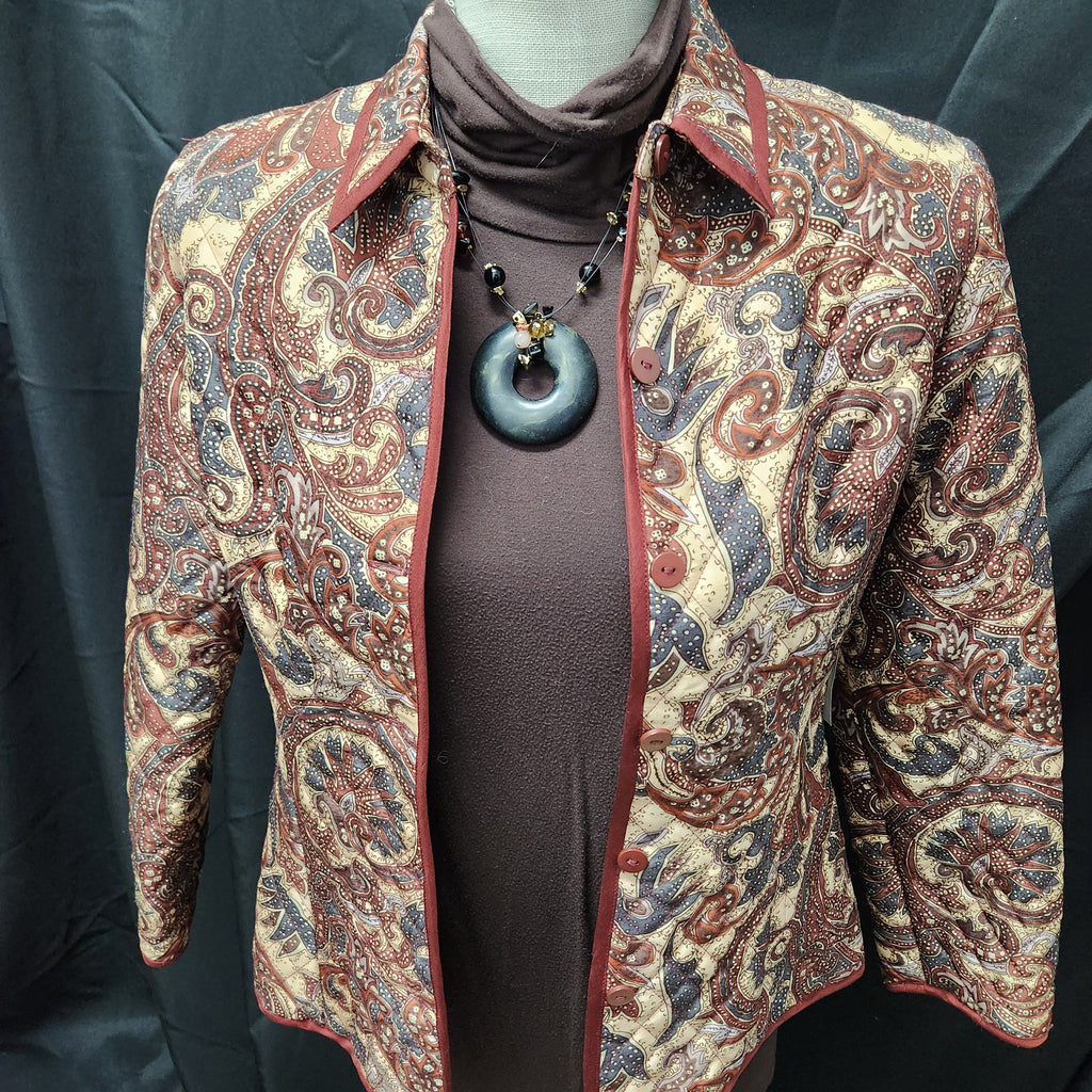 A patterned quilted Silkland beige paisley jacket , worn over a brown turtleneck sweater and styled with a large blue pendant necklace on a mannequin.