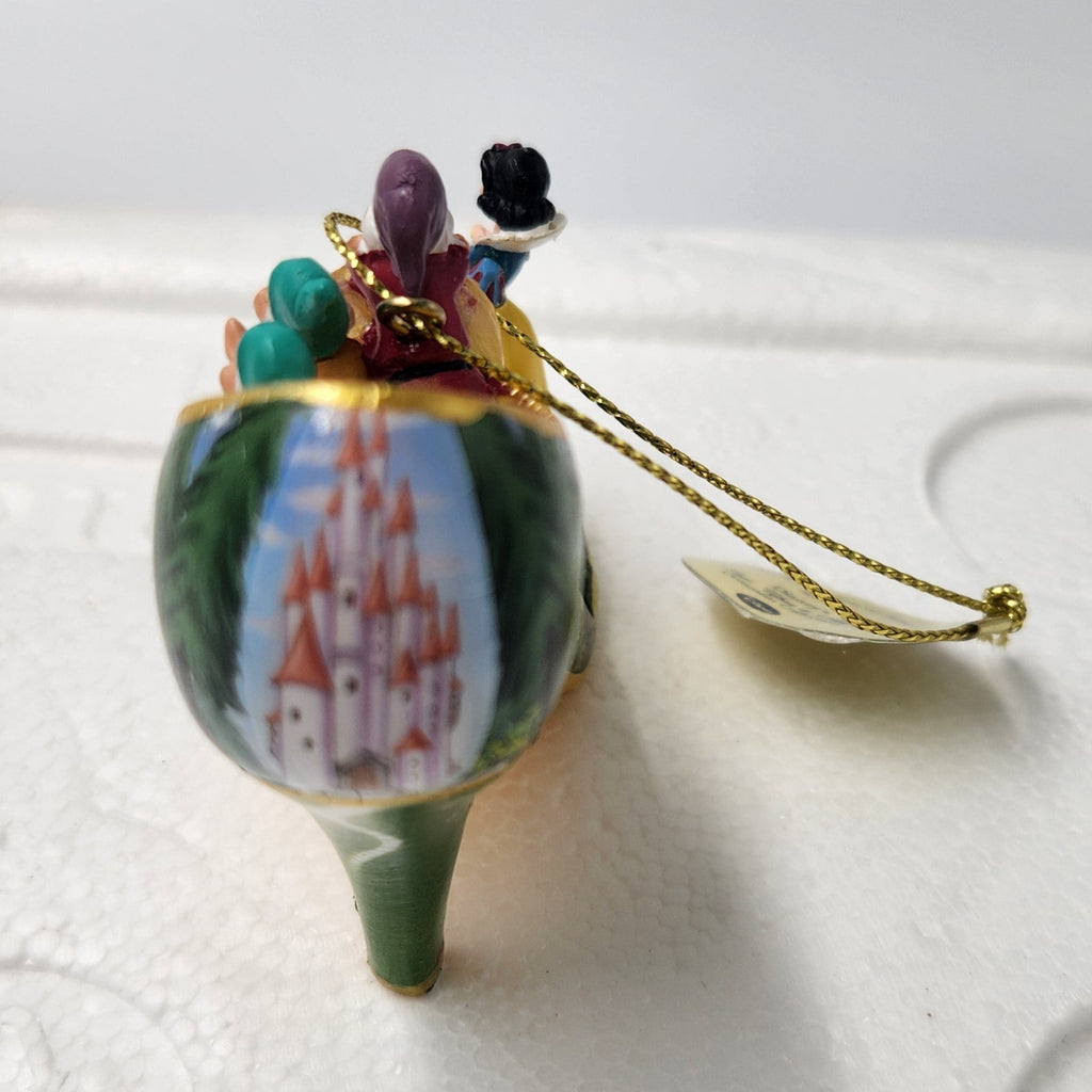 A small decorative ornament or Snow White figurine with a green stem-like base, a painted castle scene on the body, and tiny colorful details on top, resting on a white foam block with a gold hanging string.