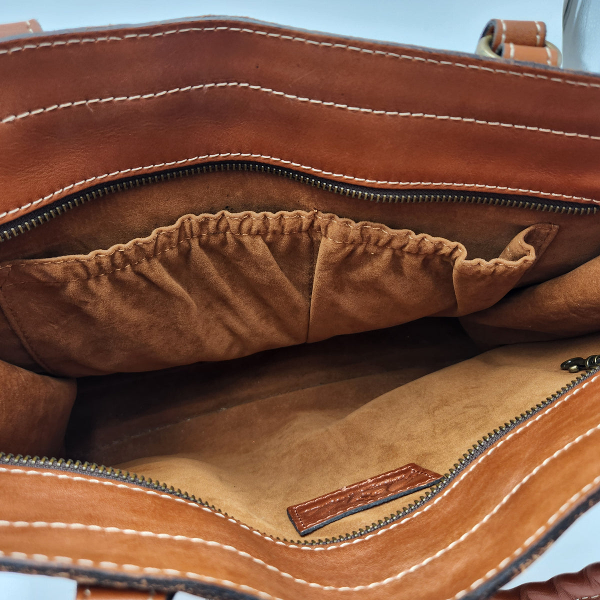 Close-up of the interior of a brown leather handbag, showing a tan suede-like lining, elastic pocket, zipper compartments, and white contrast stitching. Patricia Nash