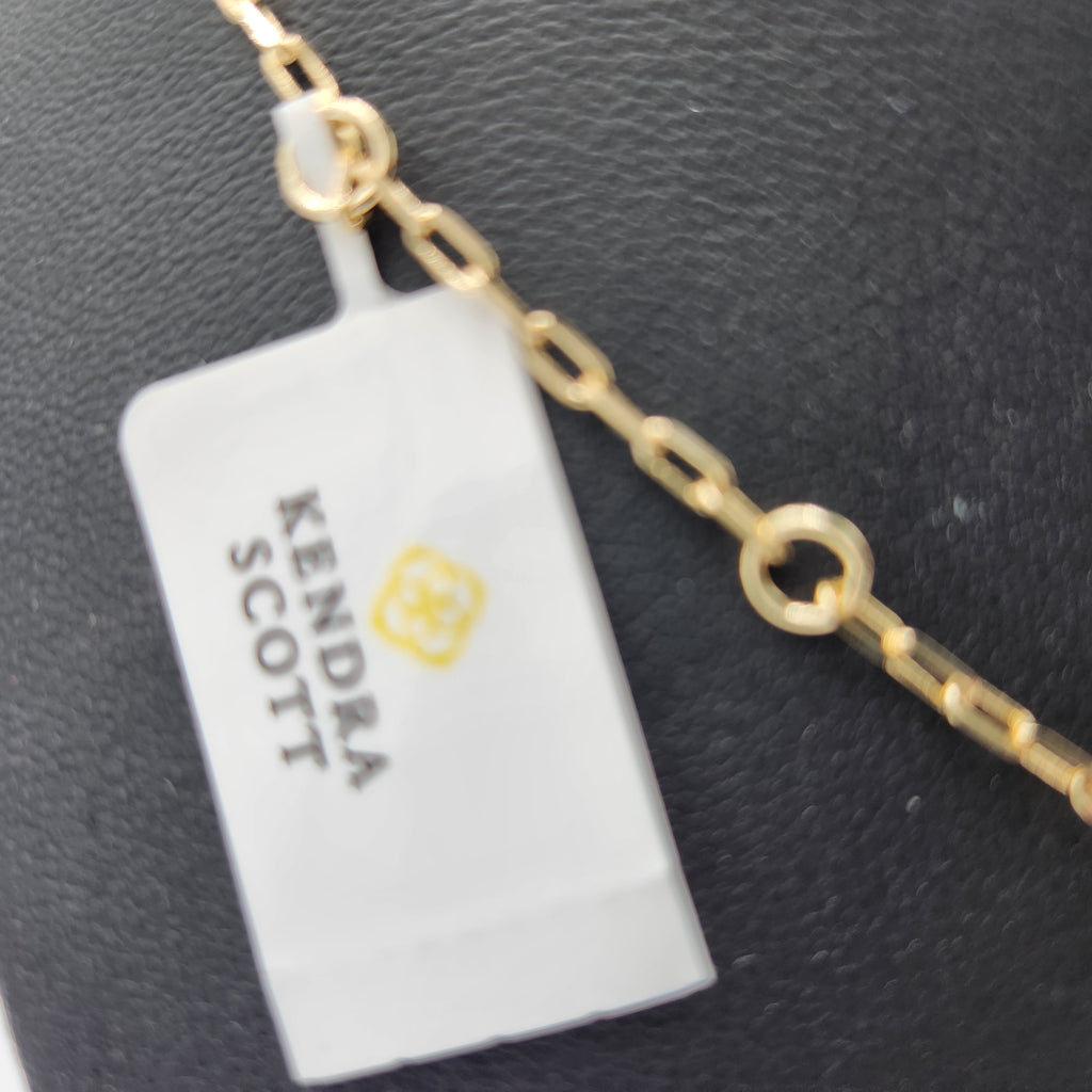 A close-up of a gold chain necklace with a rectangular pendant wrapped in a white protective plastic sleeve. The sleeve is printed with the brand name 'KENDRA SCOTT' and a small yellow floral logo, set against a dark textured background.