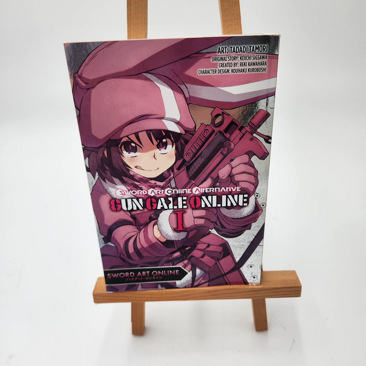 A physical copy of the Sword Art Online: Gun Gale Online Volume 1 manga book, displayed upright on a small wooden miniature easel against a plain white background.