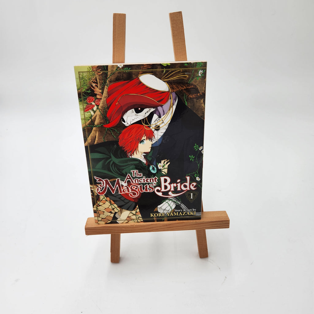 A manga book titled 'All About Magus' Bride Volume 1' displayed upright on a small wooden miniature easel against a plain white background.