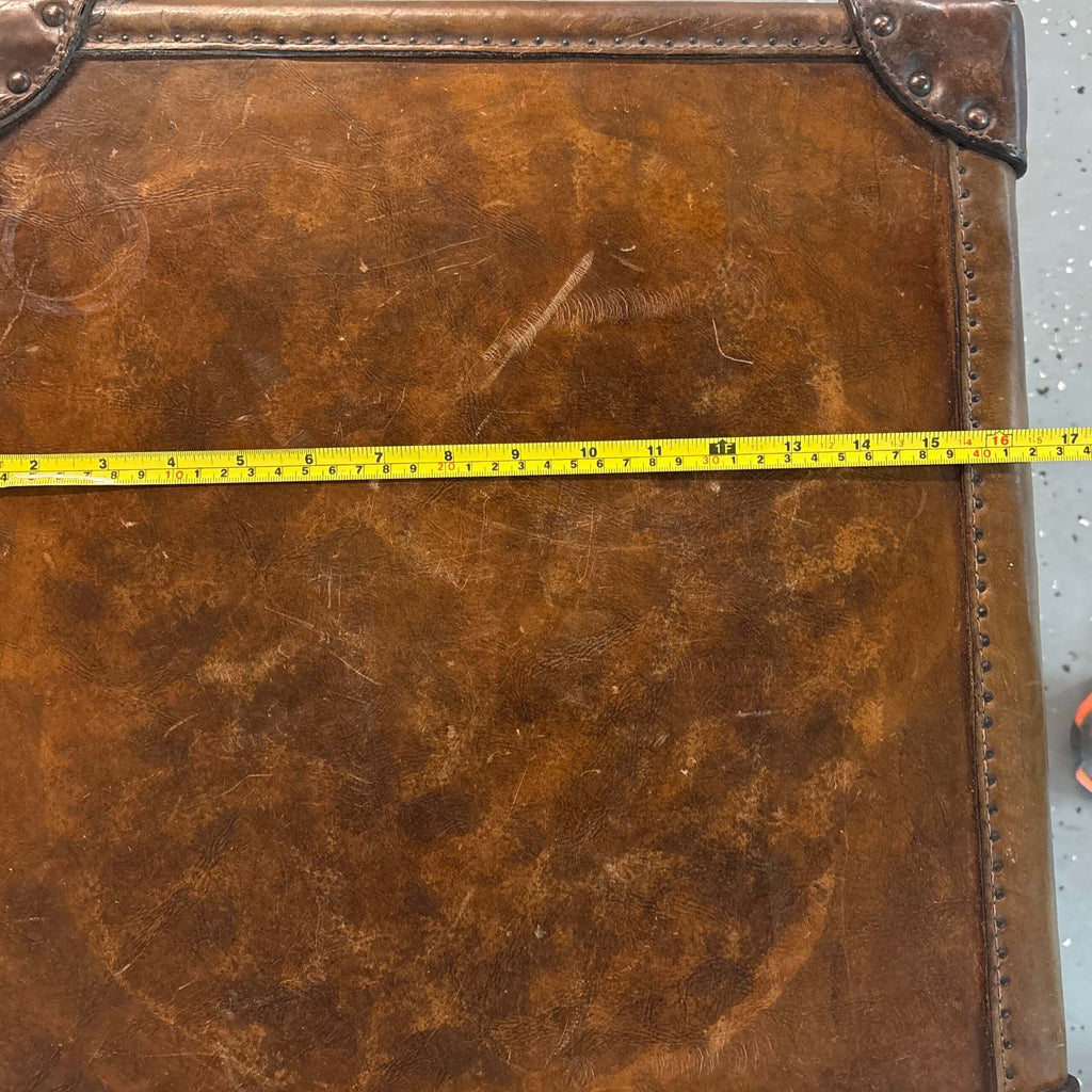 A square brown antique leather trunk top with distressed vintage styling, corner protectors, and a yellow tape measure stretched horizontally across the front to show dimensions, set on a speckled gray floor.