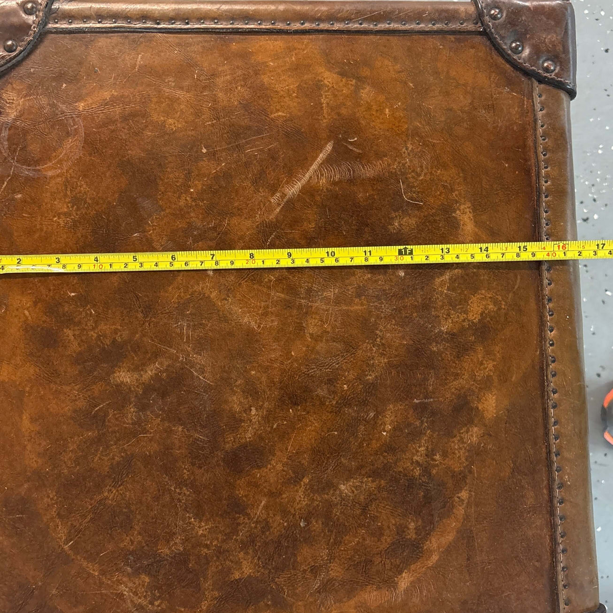 A square brown antique leather trunk top with distressed vintage styling, corner protectors, and a yellow tape measure stretched horizontally across the front to show dimensions, set on a speckled gray floor.